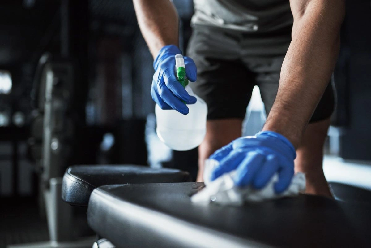 A person cleaning a gym bench with a spray bottle and cloth, wearing gloves.